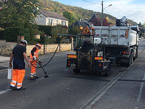 Deux ouvriers travaillent à la refection de la voirie derrière un camion transportant le mélange de bitume et d'elastomère 